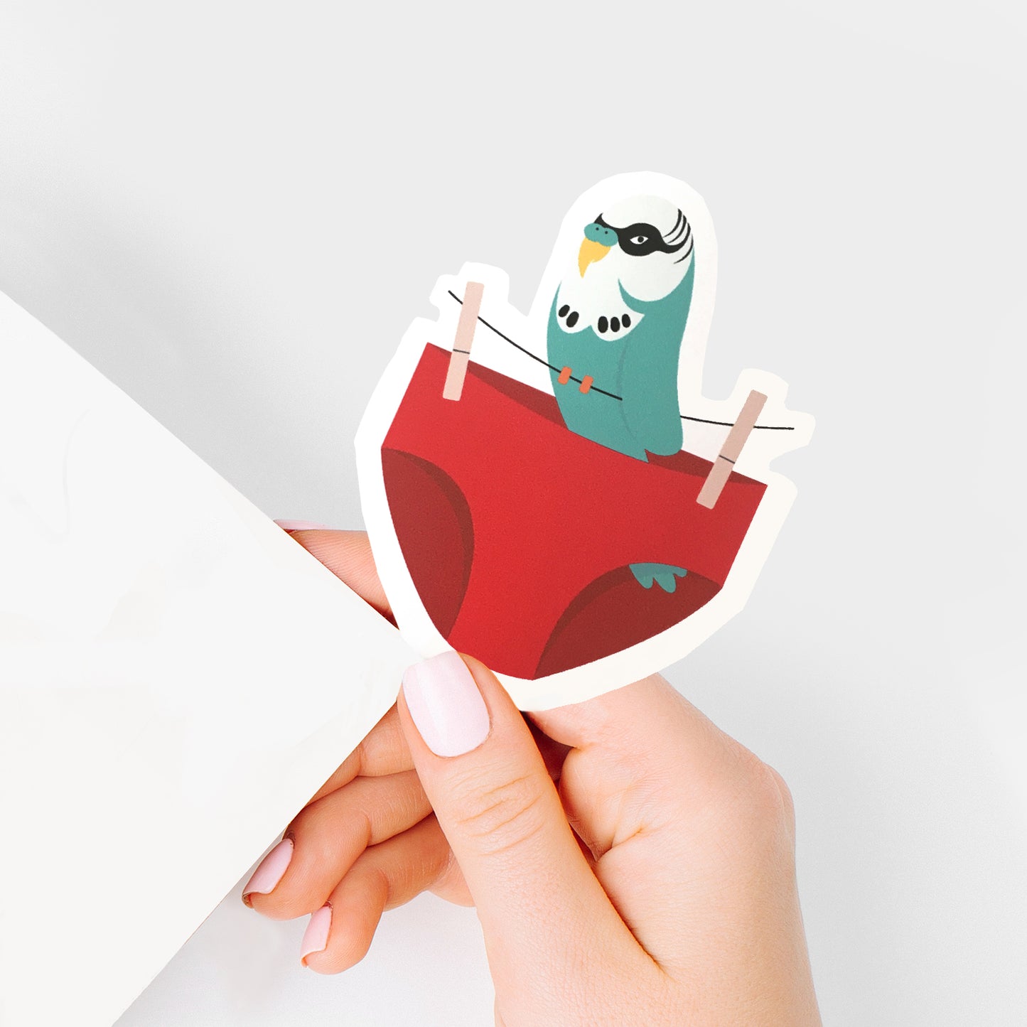 Hand holding a sticker of a budgie sitting on a washing line with pair of red budgie smugglers