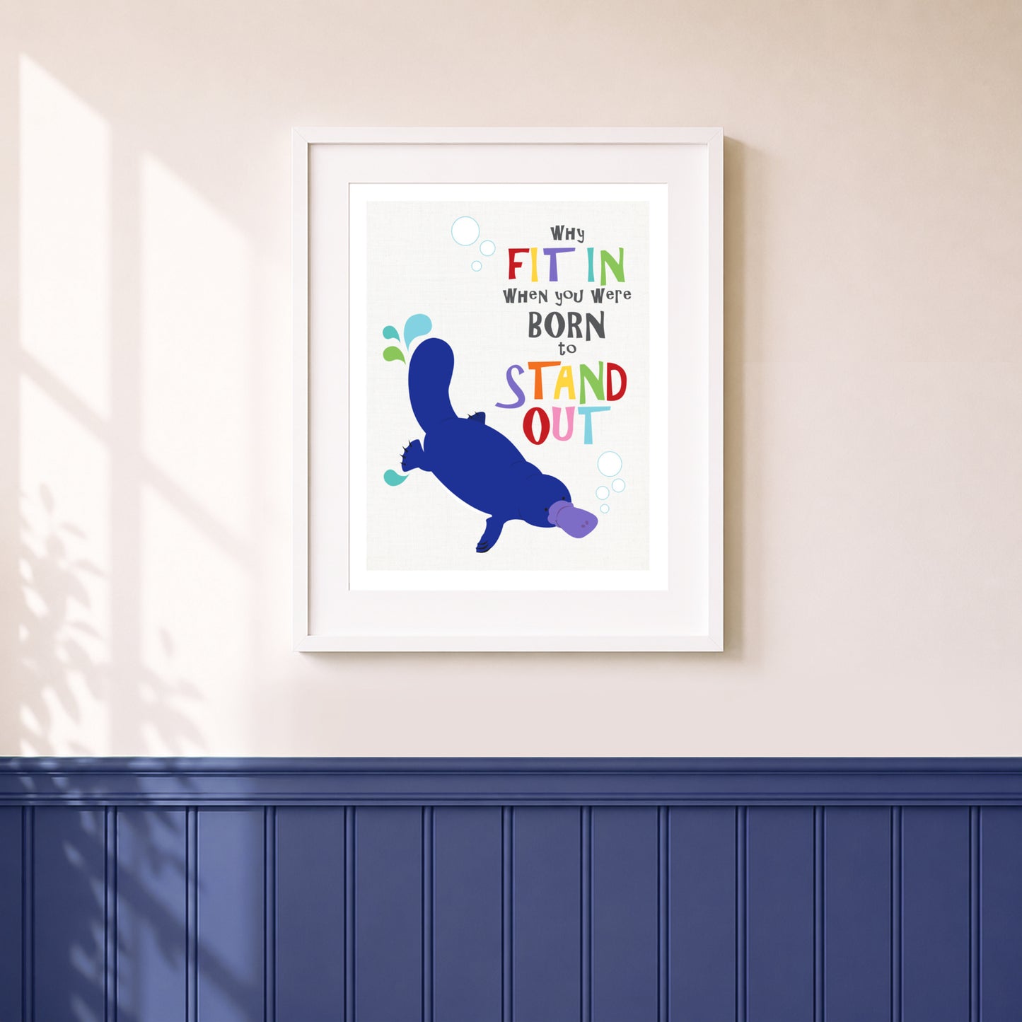 A wall art print featuring a playful platypus illustration and the phrase “WHY FIT IN When you were BORN TO STAND OUT” in colorful lettering, displayed on a wall above a navy wainscoted panel.