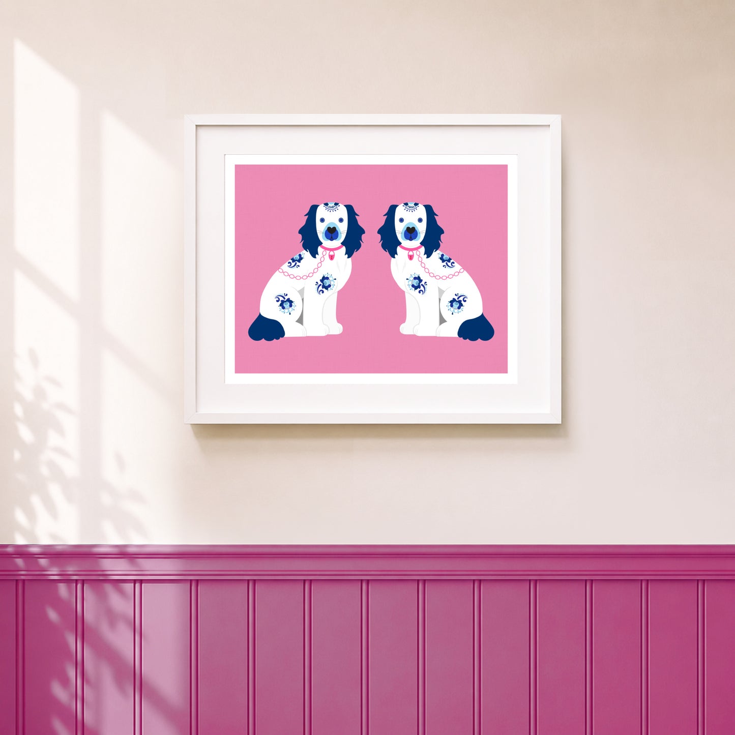 A framed art print featuring two Staffordshire spaniel dogs in white and navy with blue flower patterns, set against a solid pink background, hanging on a beige wall above a magenta wainscoted panel.