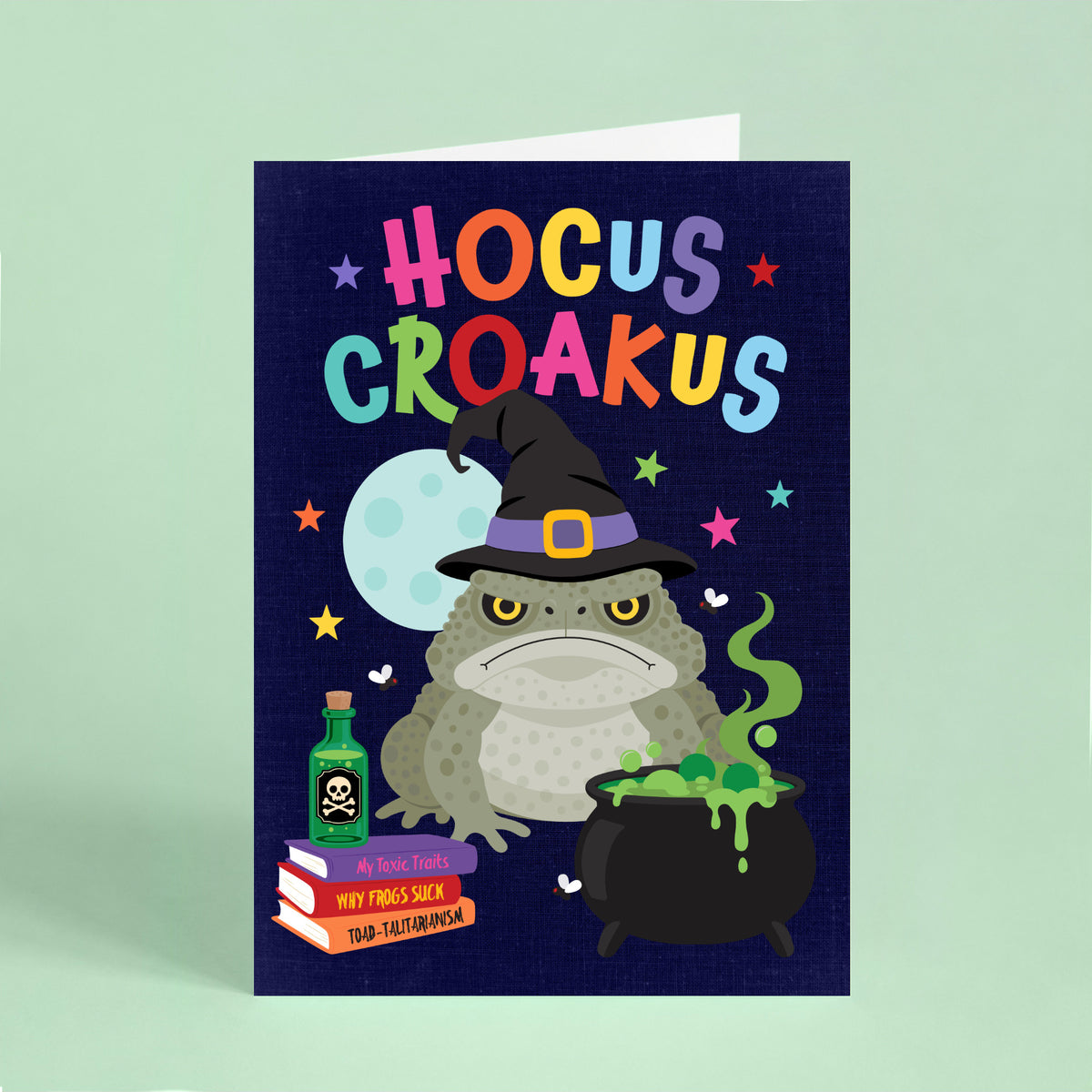 Funny Halloween card featuring a grumpy cane toad wearing a witch's hat beside a bubbling cauldron with the words "Hocus Croakus".