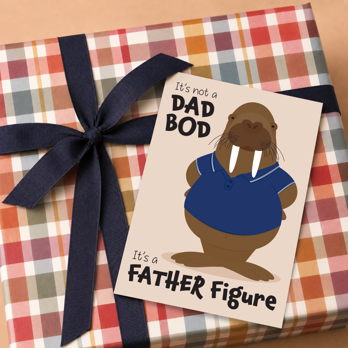 Walrus Father's Day card with the phrase "It's not a Dad Bod, It's a Father Figure" resting on a wrapped gift with colourful plaid wrapping paper and navy ribbon, funny dad gift idea.