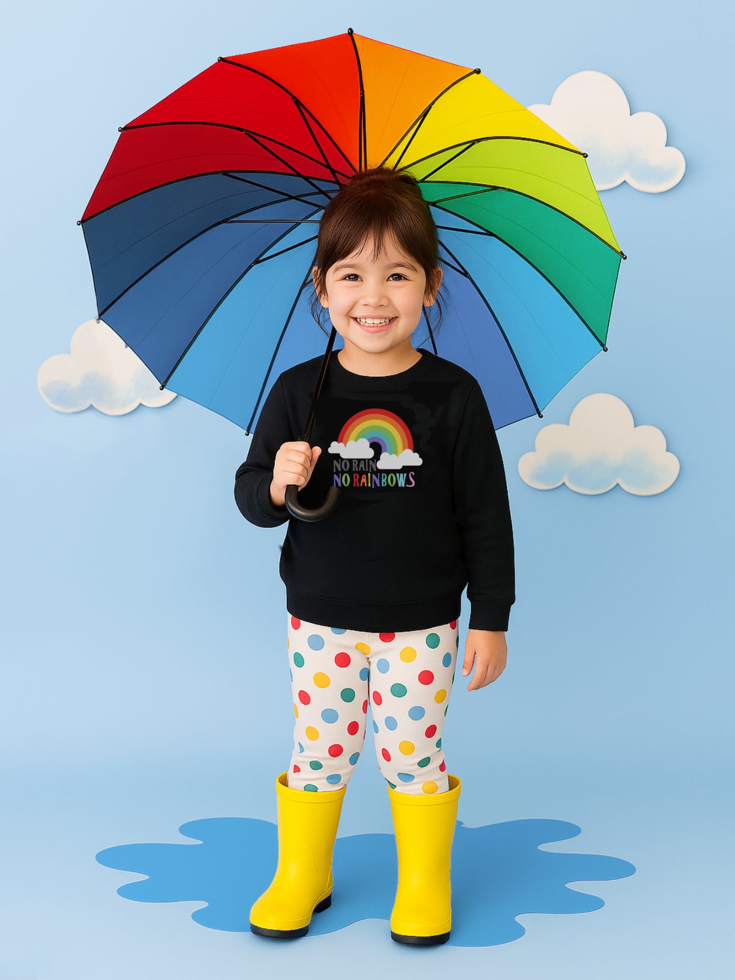 A young girl wearing a black jumper with colourful rainbow print on it and words no run no rainbows, and colourful spotty leggings stands in front of a light blue background. P