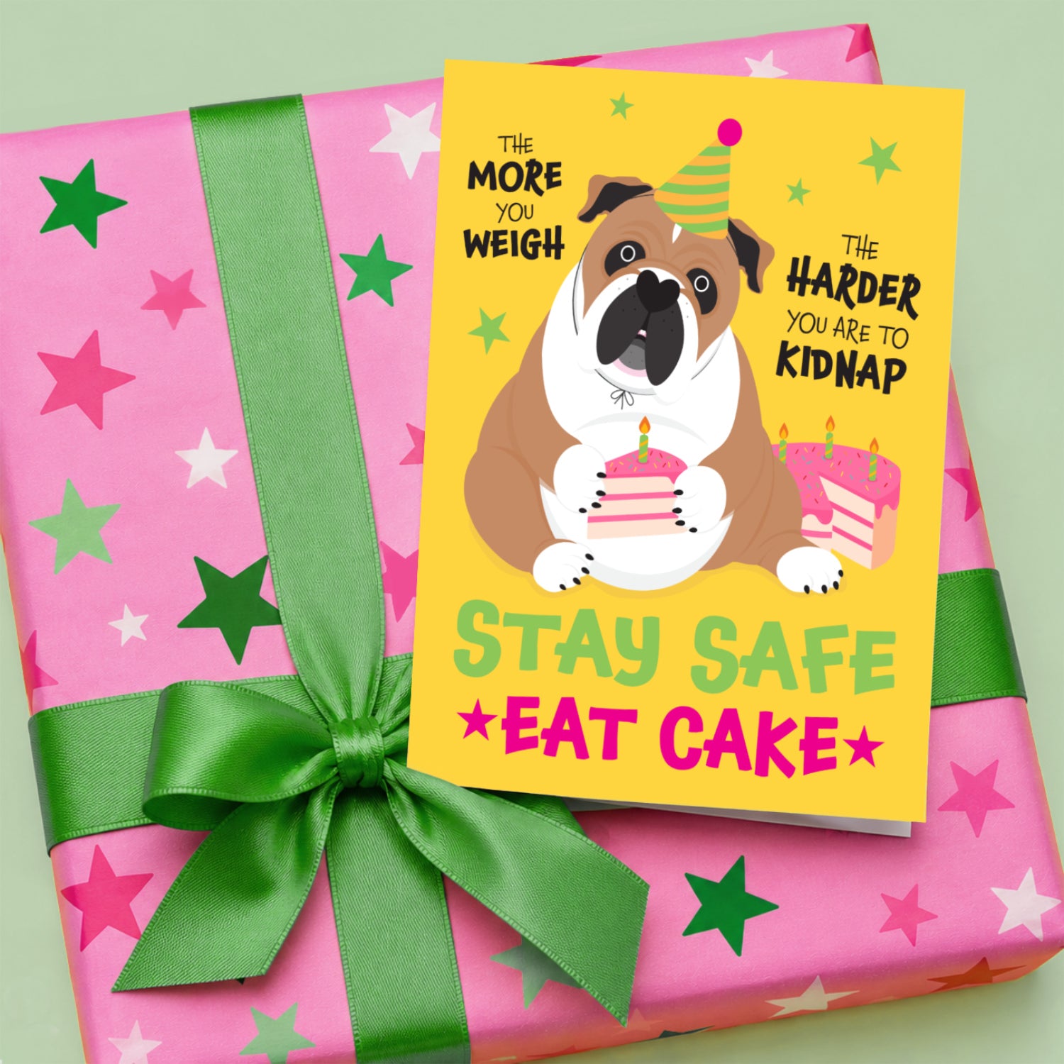 Funny English Bulldog birthday card with a colourful dog holding a slice of cake, displayed on a pink star patterned gift with a bright green ribbon and bow.