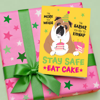 Funny English Bulldog birthday card with a colourful dog holding a slice of cake, displayed on a pink star patterned gift with a bright green ribbon and bow.
