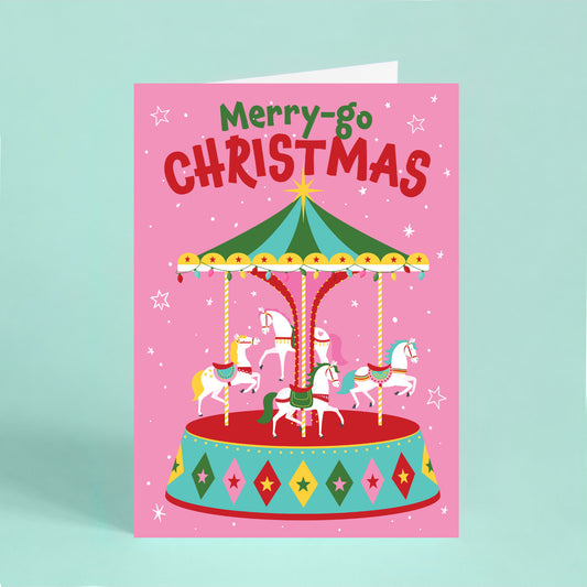 Colourful Christmas card featuring a festive carousel with white horses, Christmas lights and the words “Merry-go Christmas” on a pink background.