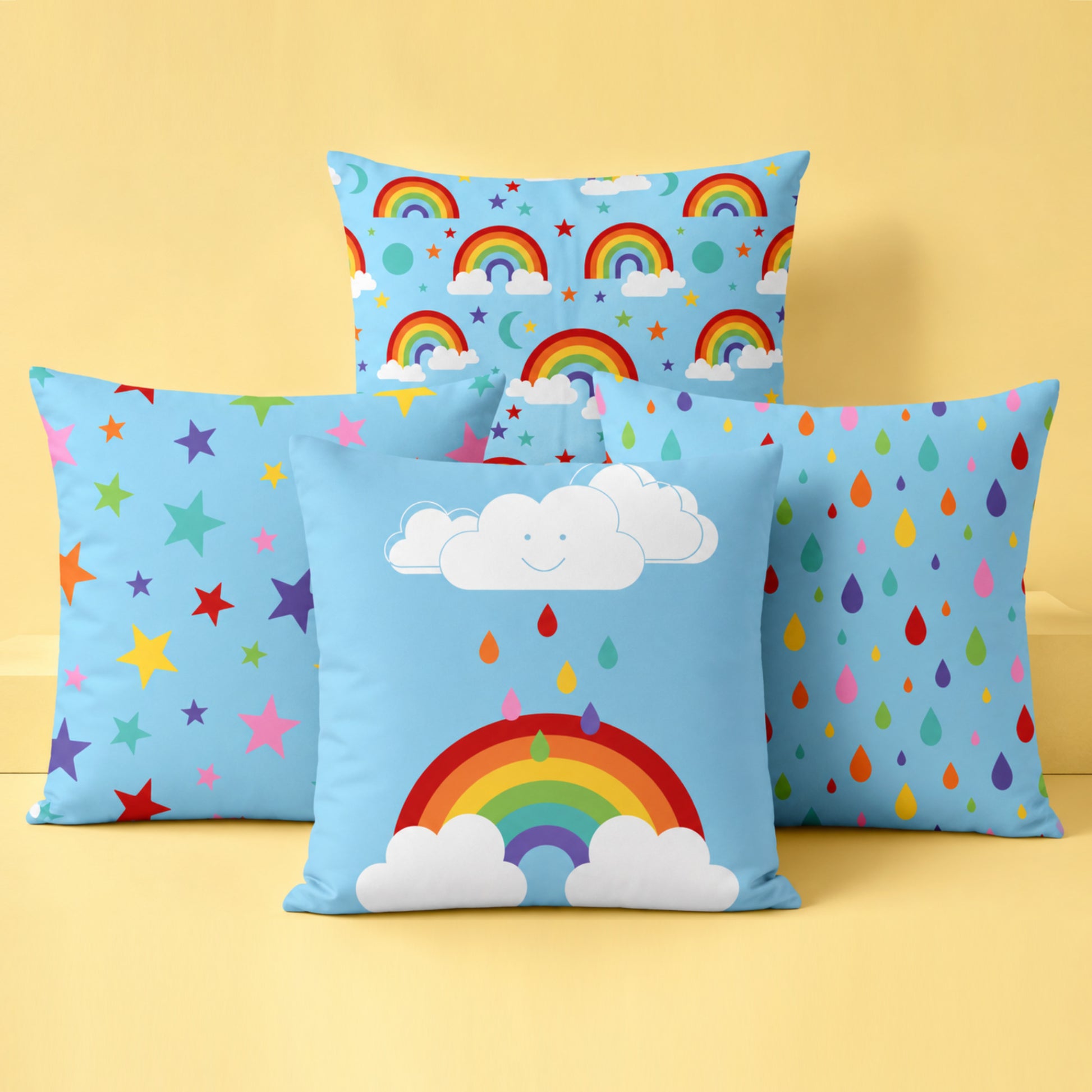 Four colourful decorative cushion covers together in a bright studio setting on a soft pastel yellow background. A rainbow and smiling cloud cushion sits at the front, with a multicoloured stars cushion and rainbow raindrops cushion positioned behind on either side. A rainbow skies pattern cushion stands at the back. The cushions feature cheerful rainbow colours on light blue fabric