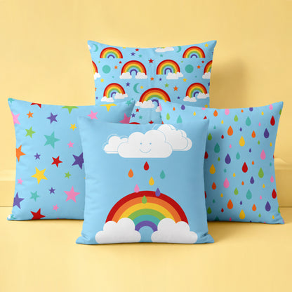 Four colourful decorative cushion covers together in a bright studio setting on a soft pastel yellow background. A rainbow and smiling cloud cushion sits at the front, with a multicoloured stars cushion and rainbow raindrops cushion positioned behind on either side. A rainbow skies pattern cushion stands at the back. The cushions feature cheerful rainbow colours on light blue fabric