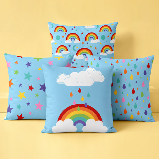 Four colourful decorative cushion covers together in a bright studio setting on a soft pastel yellow background. A rainbow and smiling cloud cushion sits at the front, with a multicoloured stars cushion and rainbow raindrops cushion positioned behind on either side. A rainbow skies pattern cushion stands at the back. The cushions feature cheerful rainbow colours on light blue fabric