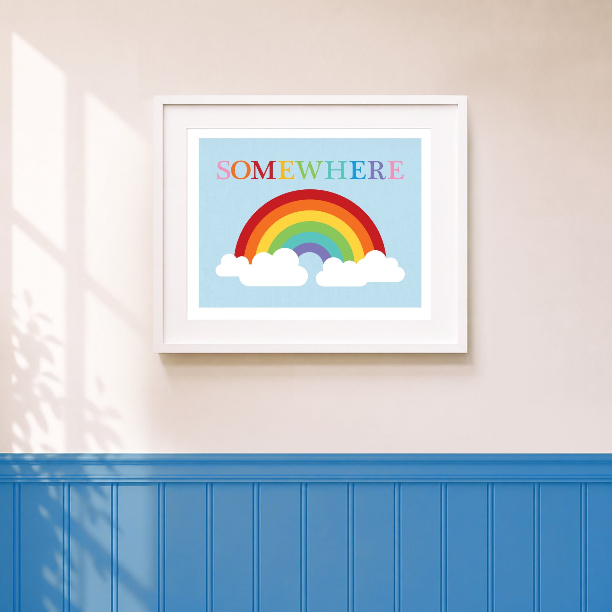 An art print featuring a colorful rainbow and the word 'SOMEWHERE' in colourful letters, displayed on a wall above blue wainscoting.