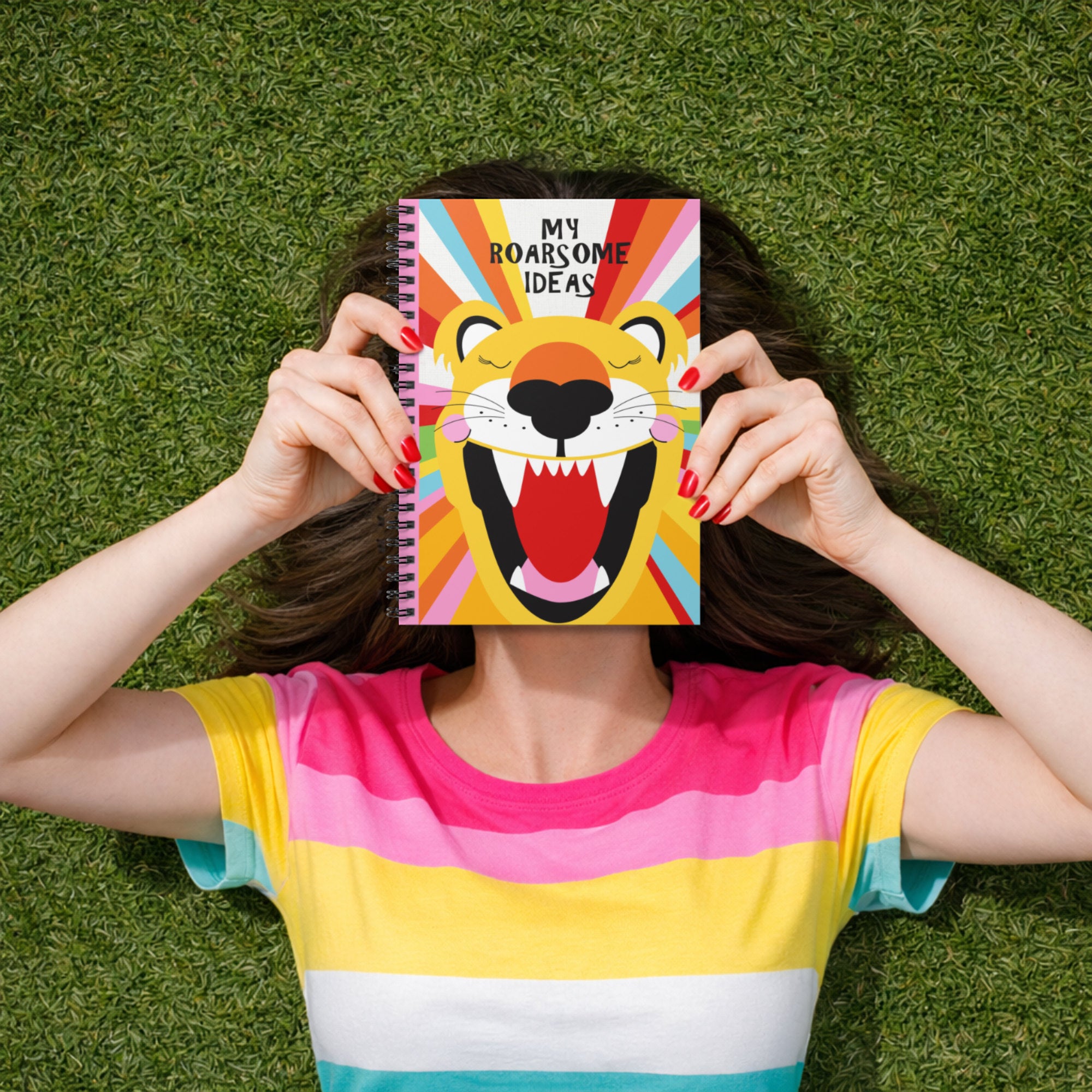 Woman lying on green grass holding a colourful spiral notebook with a roaring lion illustration that reads “My Roarsome Ideas,” wearing a bright rainbow striped top