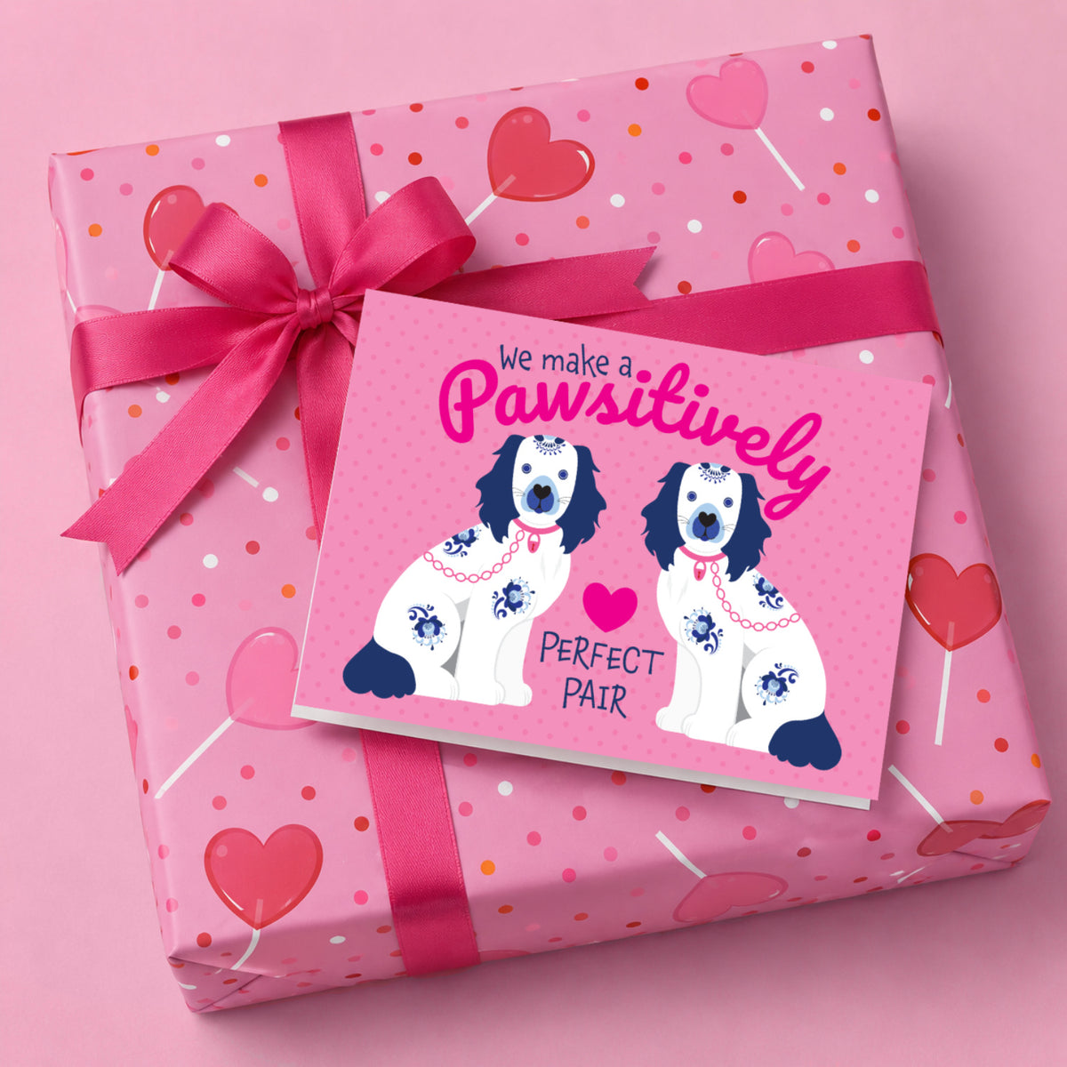 A pink gift-wrapped package tied with a matching pink ribbon and bow, with a  greeting card placed on top. The card features an illustration of two Staffordshire ceramic dogs in blue and white with pink collars, and the text 'We make a Pawsitively PERFECT PAIR'.