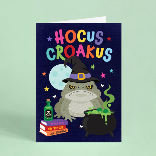 Funny Halloween card featuring a grumpy cane toad wearing a witch's hat beside a bubbling cauldron with the words "Hocus Croakus".
