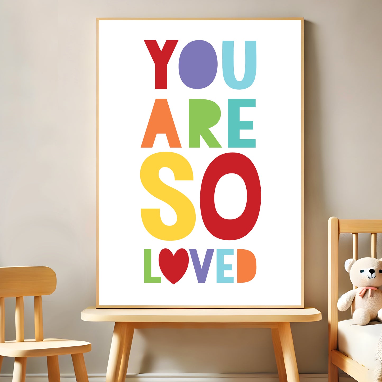 A colourful nursery print with the phrase "You Are So Loved" in bold rainbow-coloured letters, displayed in a minimal wooden frame on a light-toned wall. The decor includes a wooden chair and a teddy bear on a small bench.