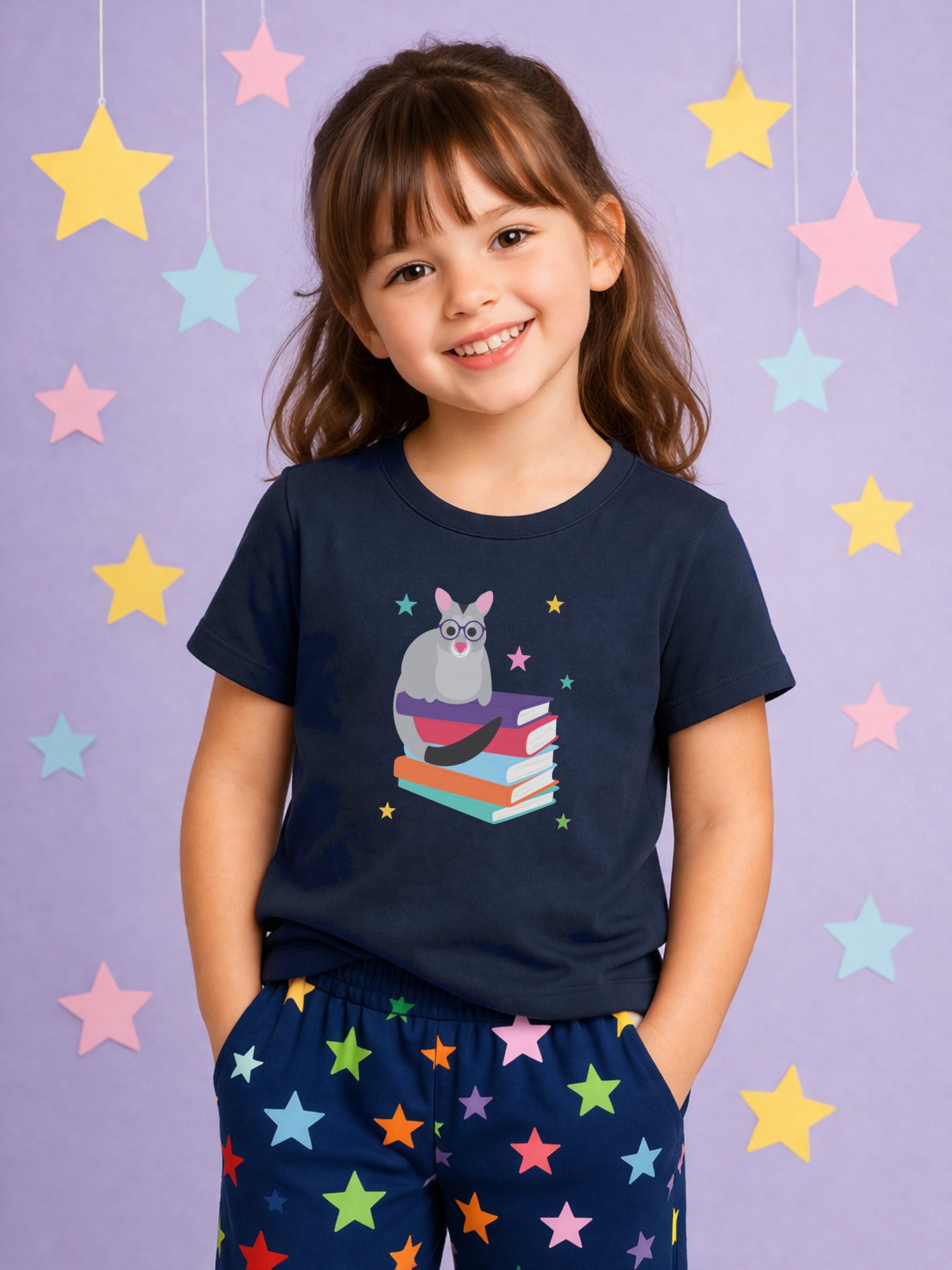 Smiling young girl wearing a navy t-shirt with possum and book design and colourful star print pajama pants, standing against a soft purple studio background with hanging pastel stars,
