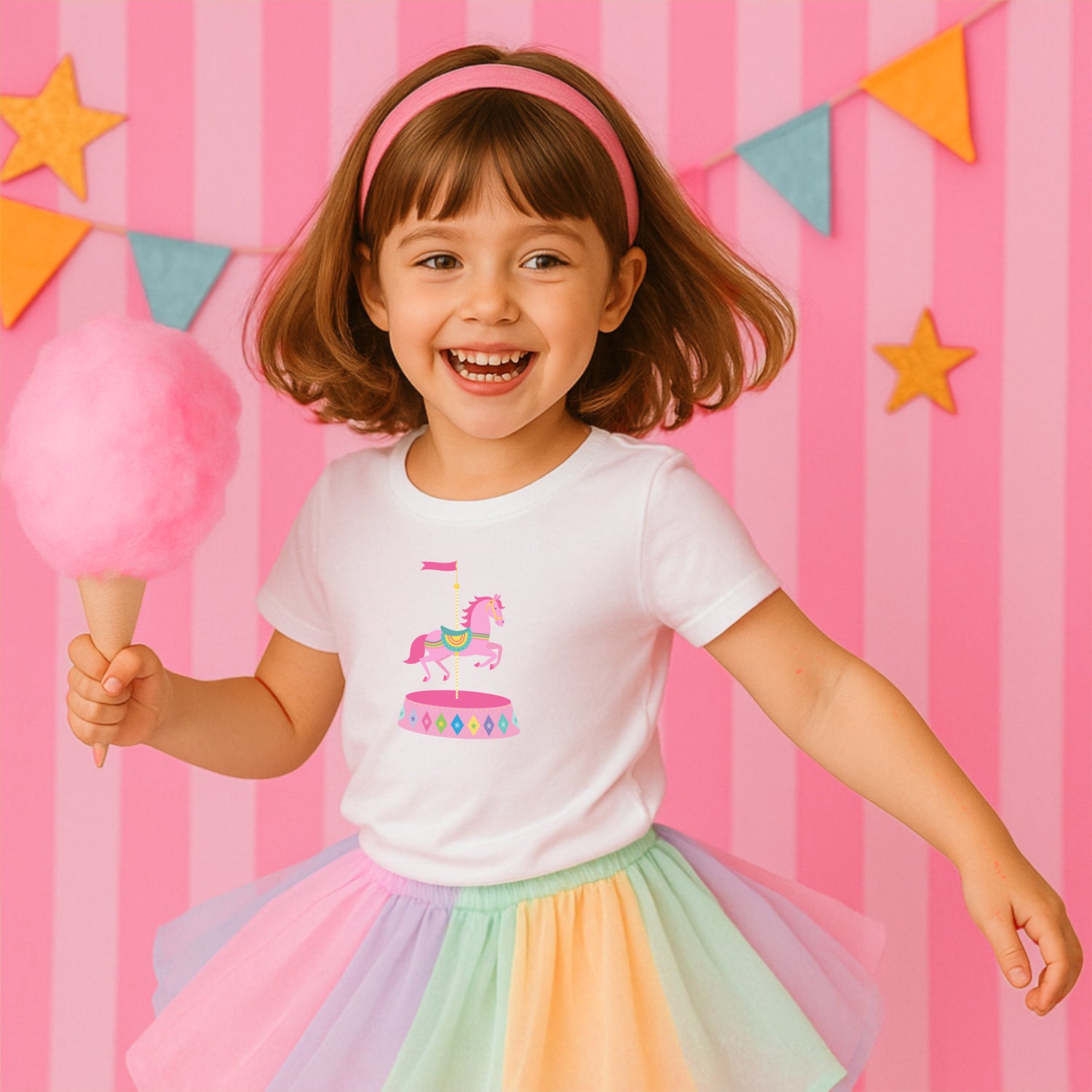 Child wearing a white t-shirt with a colorful carousel horse design, holding pink cotton candy against a pink striped background with star decorations.