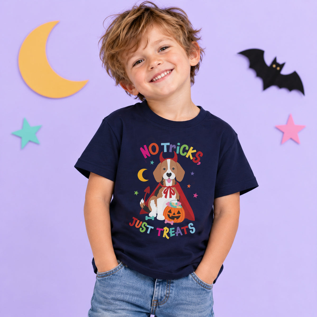 A smiling young boy wearing a navy blue graphic t-shirt with a Halloween dog design that reads 'NO TRICKS, JUST TREATS'. The background is a light purple wall decorated with paper stars, a crescent moon, and a bat.