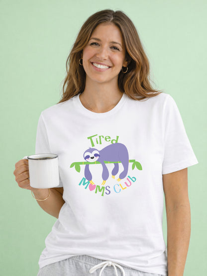 Smiling woman wearing a white Tired Mums Club t-shirt with colourful sloth design, holding a coffee against a soft green background