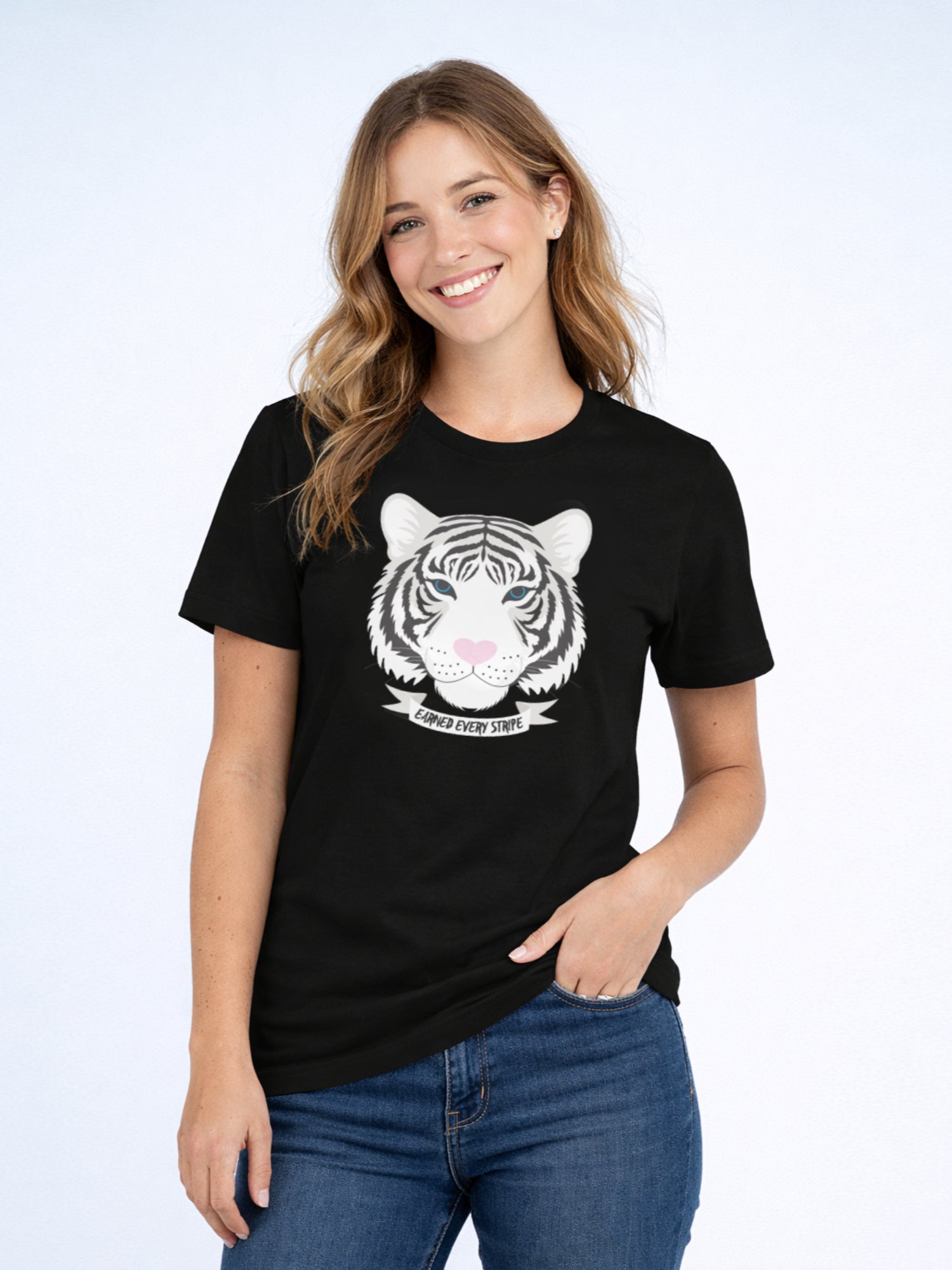 Smiling woman wearing a black t-shirt with a white tiger illustration and the phrase “Earned Every Stripe”, standing against a soft light blue background in a relaxed pose.