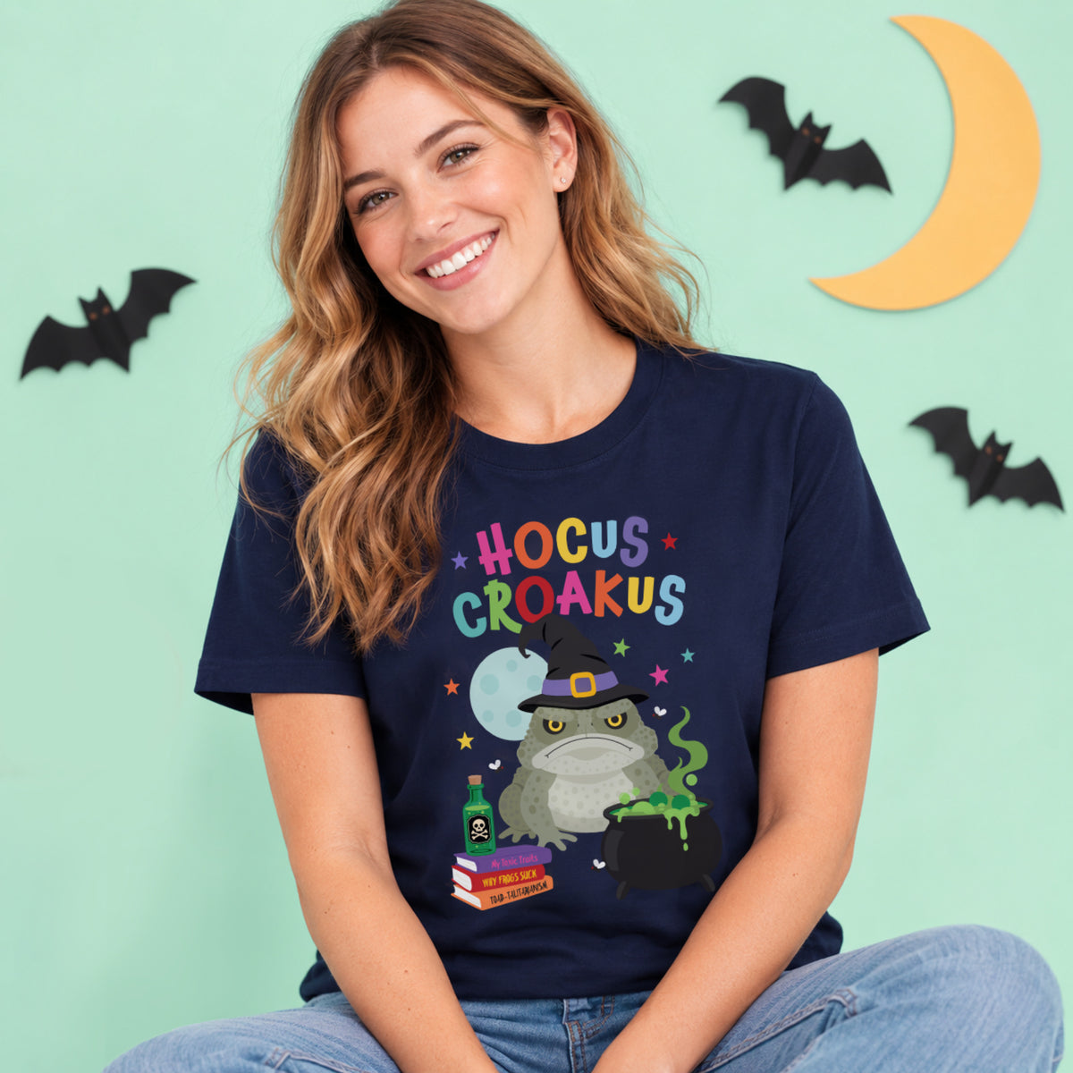 Woman wearing a navy Hocus Croakus Halloween T-shirt featuring a colourful witch toad and cauldron design, photographed against a light mint  Halloween backdrop.