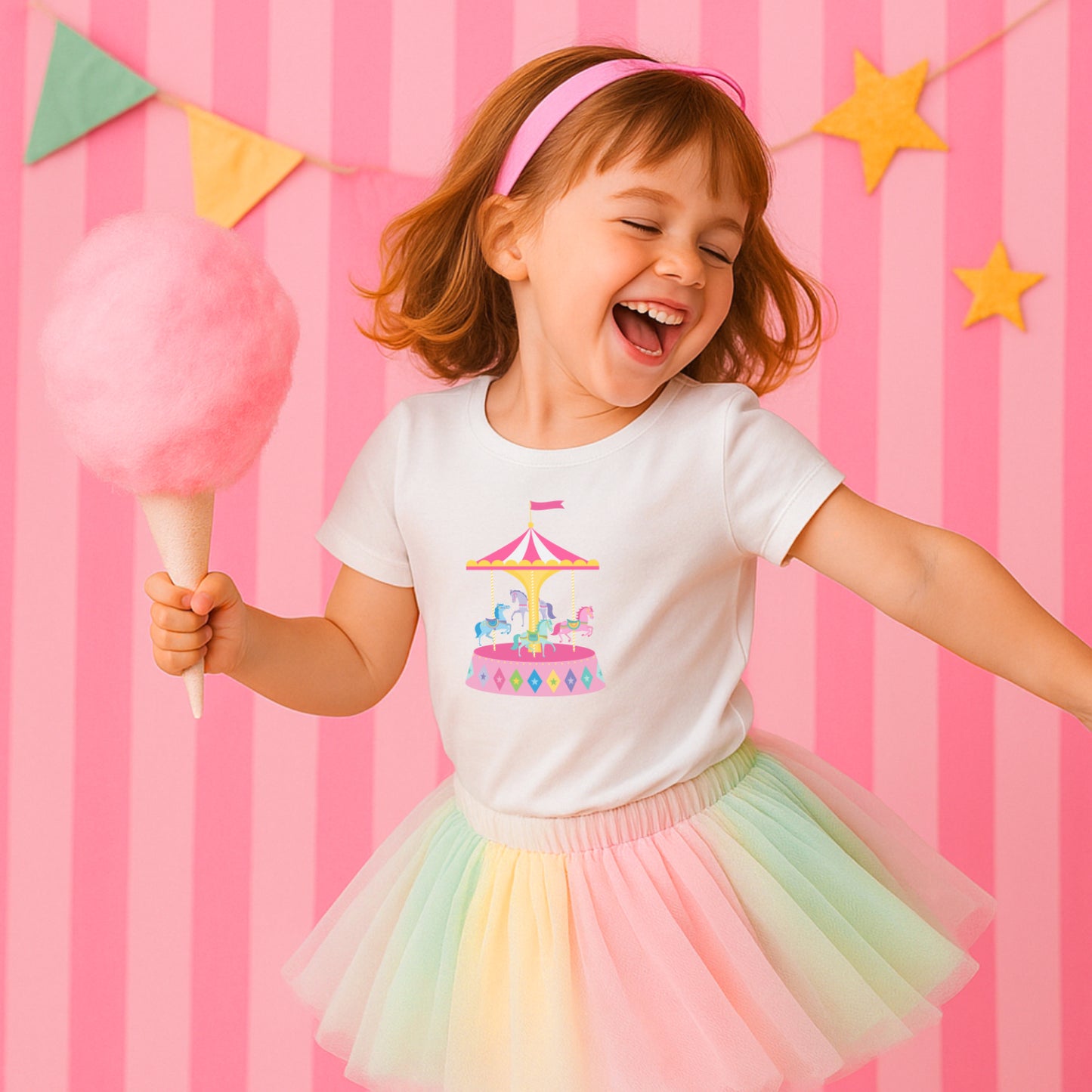 Child wearing a white t-shirt with a carousel design and a rainbow tutu skirt, holding cotton candy against a pink striped background.
