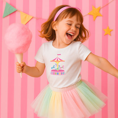 Child wearing a white t-shirt with a carousel design and a rainbow tutu skirt, holding cotton candy against a pink striped background.
