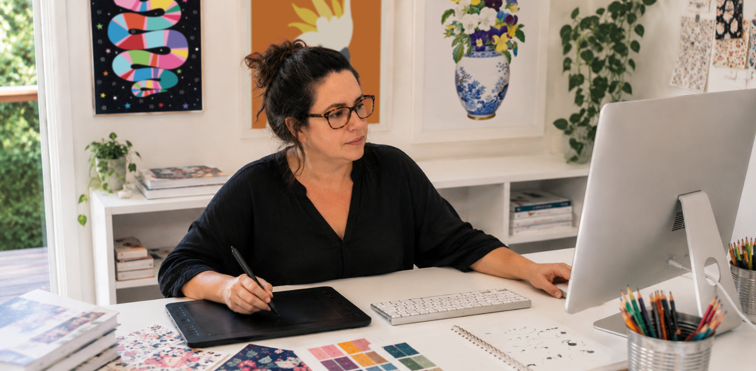 Surface pattern designer working at a desk with a computer and art supplies in a home office setting.