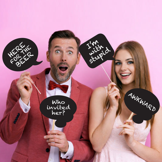 Two people holding humorous photo booth props with text on a pink background