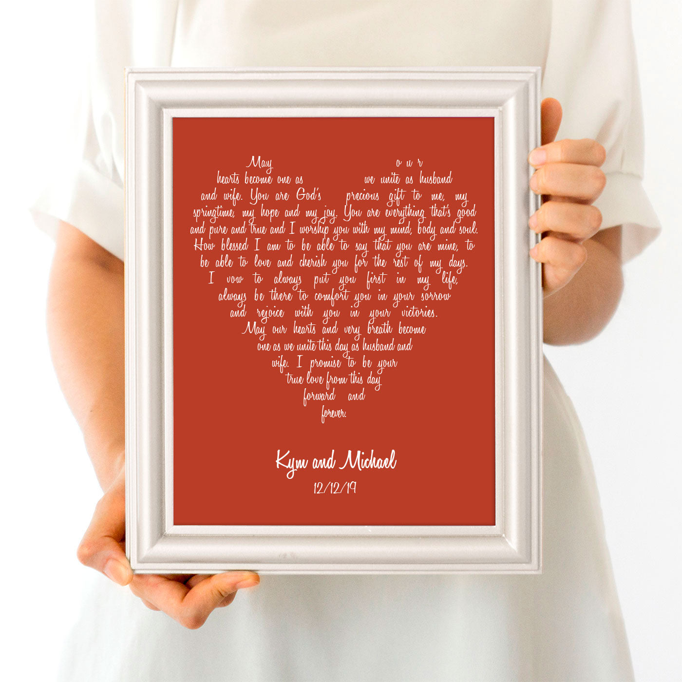 A personalized heart-shaped artwork print with wedding vows written in a mixed font, held in a hand. The background is orange, and the text is in a white heart-shaped area on the front of the picture frame.
