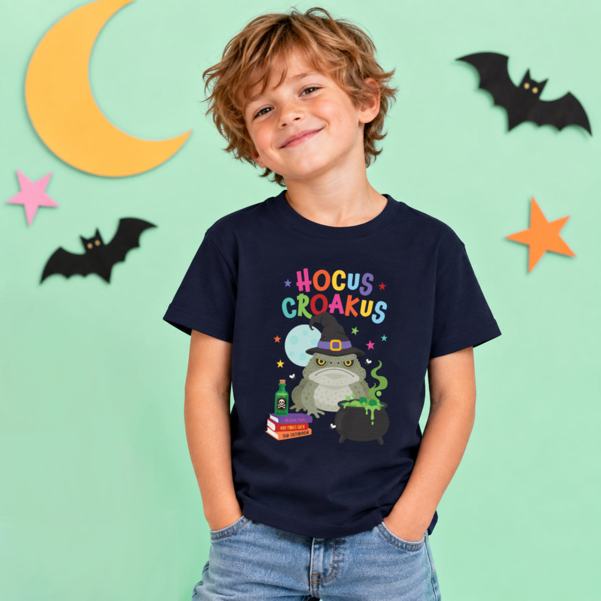 Child wearing a navy Hocus Croakus Halloween T-shirt featuring a colourful witch cane toad, bubbling cauldron and potion design against a light mint Halloween backdrop.